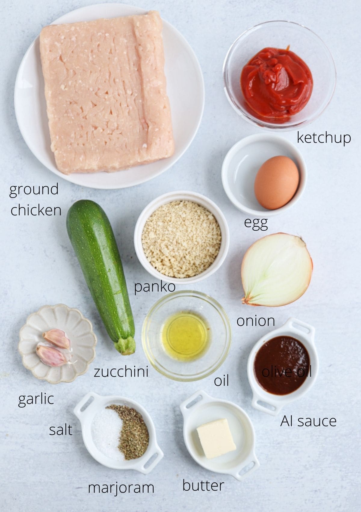 Top-down view of labeled ingredients for a chicken potato broccoli casserole: ground chicken, ketchup, egg, panko breadcrumbs, onion, zucchini, garlic, salt, marjoram, olive oil, butter, and A1 sauce on a light surface.