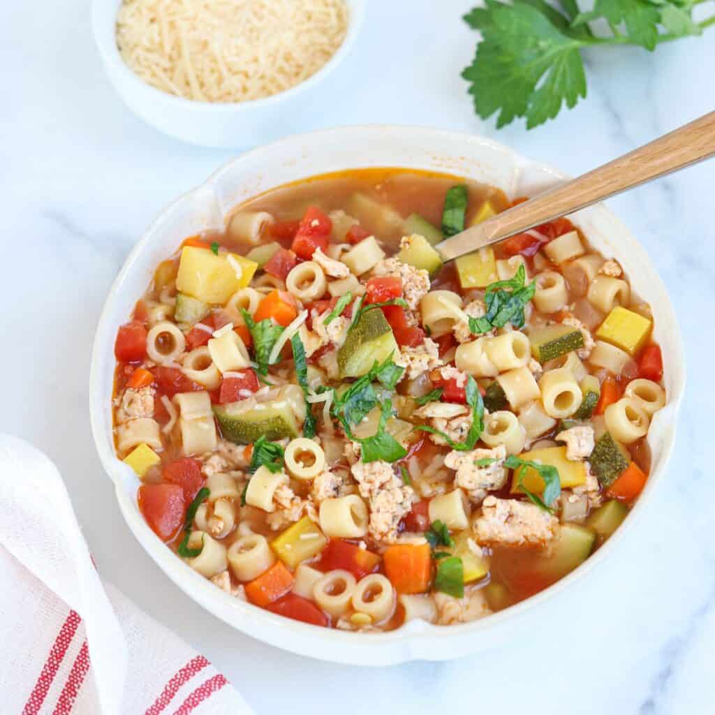 A bowl of vegetable and pasta soup with chopped zucchini, tomatoes, carrots, ditalini pasta, and herbs, garnished with fresh basil. A wooden spoon rests in the bowl. A dish of grated cheese is nearby.