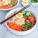 A bowl filled with brown rice, shredded carrots, ground meat, sliced cucumber, kimchi, chopped green onions, half a soft-boiled egg, and a dollop of creamy sauce. Chopsticks rest on the bowl.