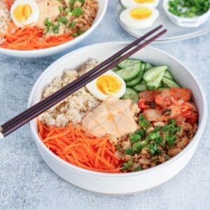 A bowl filled with brown rice, shredded carrots, ground meat, sliced cucumber, kimchi, chopped green onions, half a soft-boiled egg, and a dollop of creamy sauce. Chopsticks rest on the bowl.
