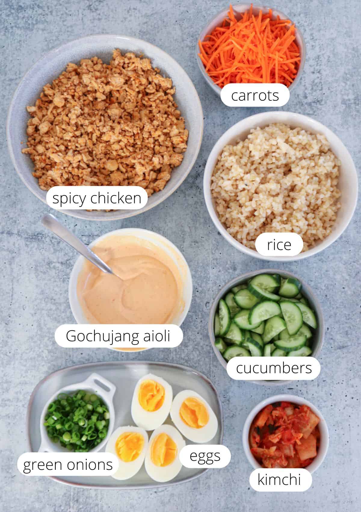 Bowls containing spicy chicken, shredded carrots, rice, sliced cucumbers, kimchi, green onions, halved boiled eggs, gochujang aioli, are arranged neatly on a gray surface, each labeled.