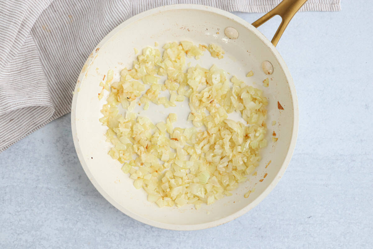 Chopped onions sautéing in a light-colored frying pan with a golden handle, ready to be added to a chicken potato broccoli casserole, placed on a light surface next to a beige striped cloth.