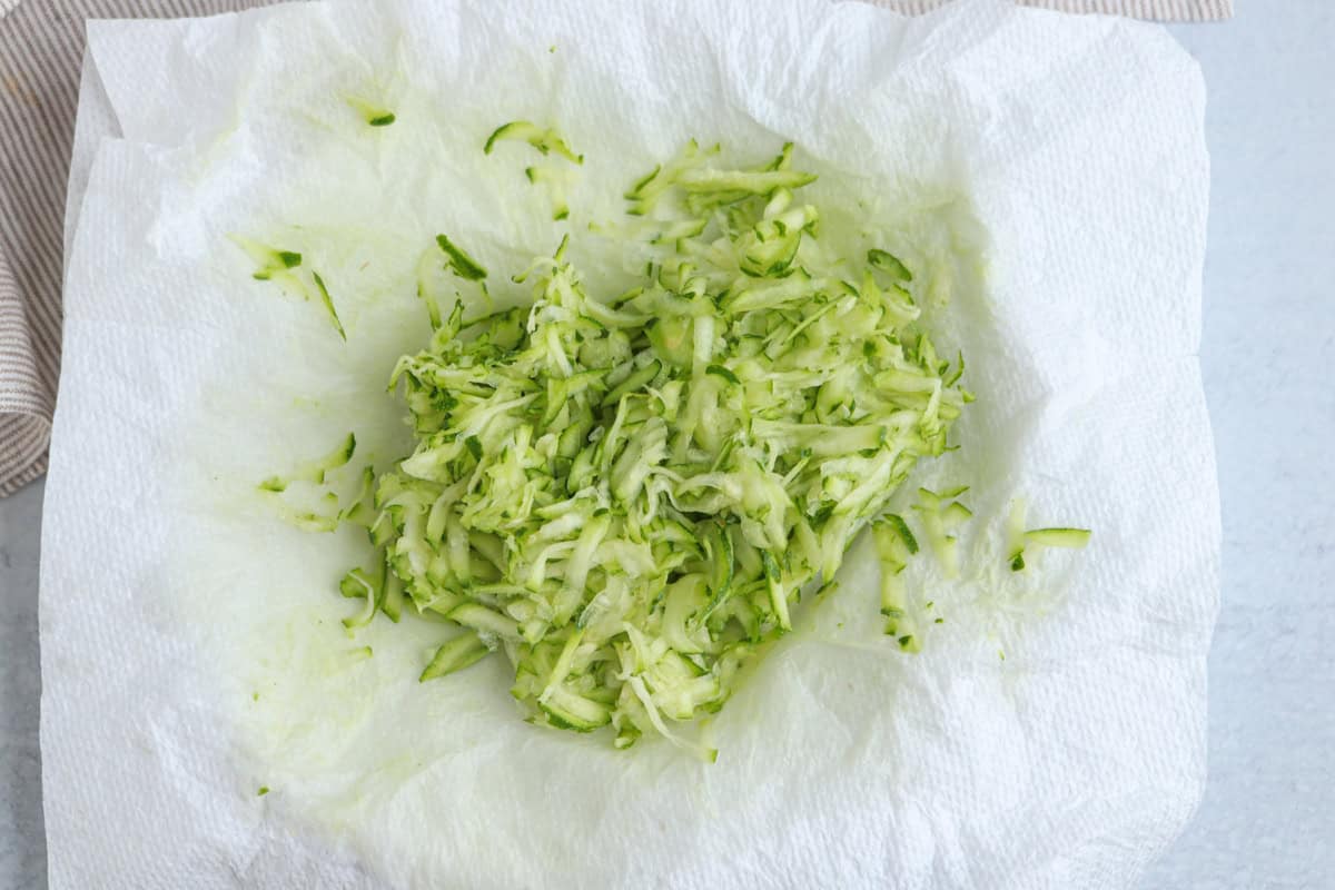 Shredded zucchini rests on a white paper towel, which is absorbing excess moisture—an essential step before adding it to dishes like chicken potato broccoli casserole. The setup is on a light surface next to beige striped fabric.