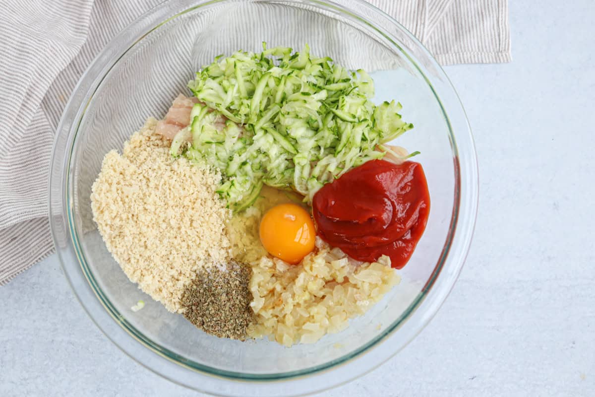 A glass bowl containing shredded zucchini, an egg, breadcrumbs, diced onions, ketchup, ground black pepper, and raw ground meat—perfect ingredients for dishes like chicken potato broccoli casserole—sits on a light surface with a striped cloth nearby.