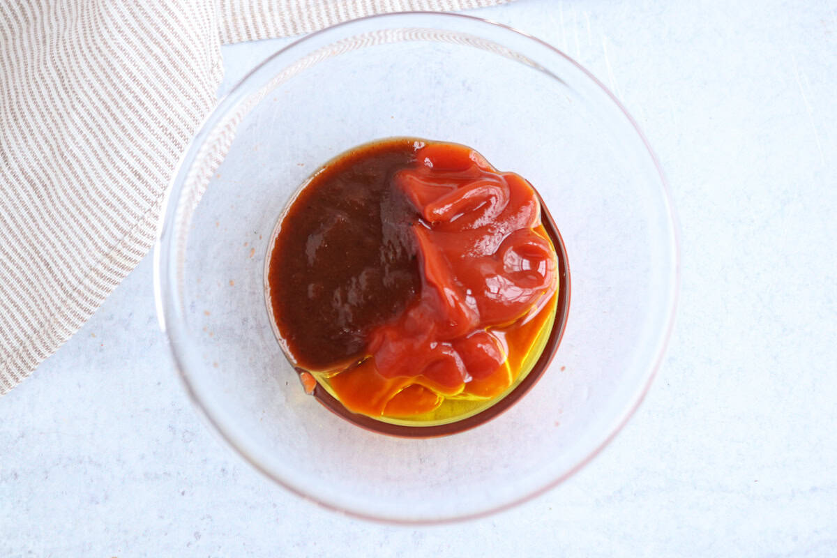 A clear glass bowl containing three sauces—dark brown, red, and yellow—placed on a light surface next to a beige and white striped cloth, perfect for serving with chicken potato broccoli casserole.