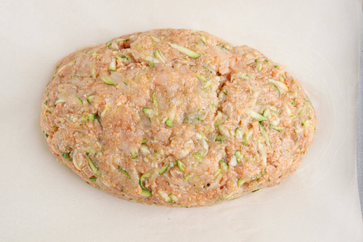 Uncooked meatloaf mixture shaped into an oval, containing ground meat and shredded vegetables like in a chicken potato broccoli casserole, resting on a sheet of parchment paper.
