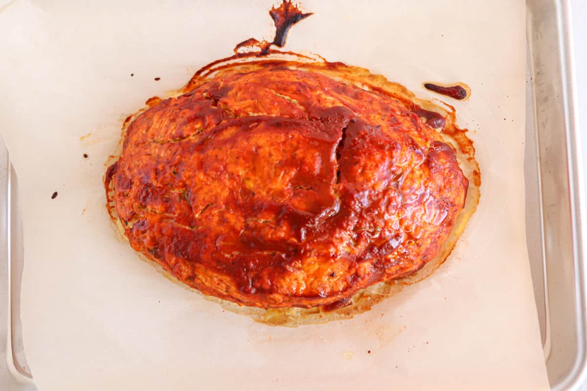 A baked meatloaf with a shiny, caramelized glaze sits on parchment paper atop a baking sheet. Its browned, slightly cracked surface is reminiscent of a chicken potato broccoli casserole, with sauce puddled around the edges.