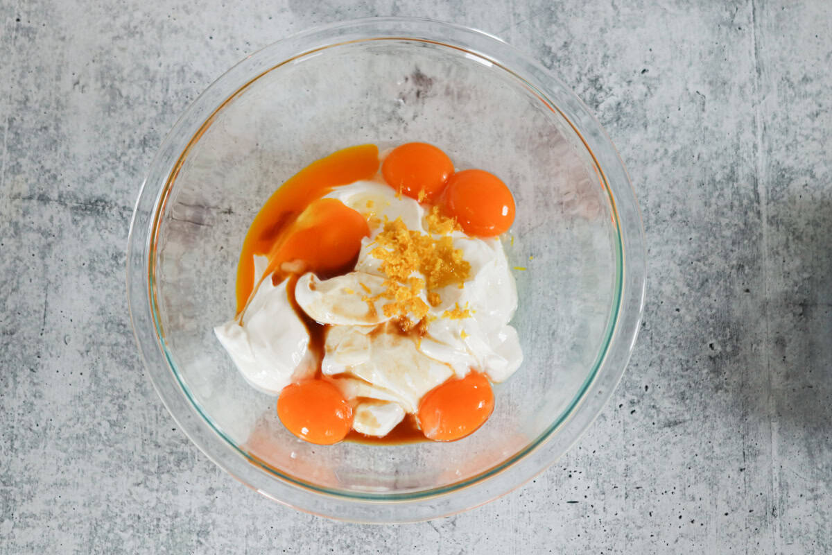 A glass bowl on a gray countertop containing four raw eggs, a dollop of white yogurt or sour cream, vanilla extract, and lemon zest.