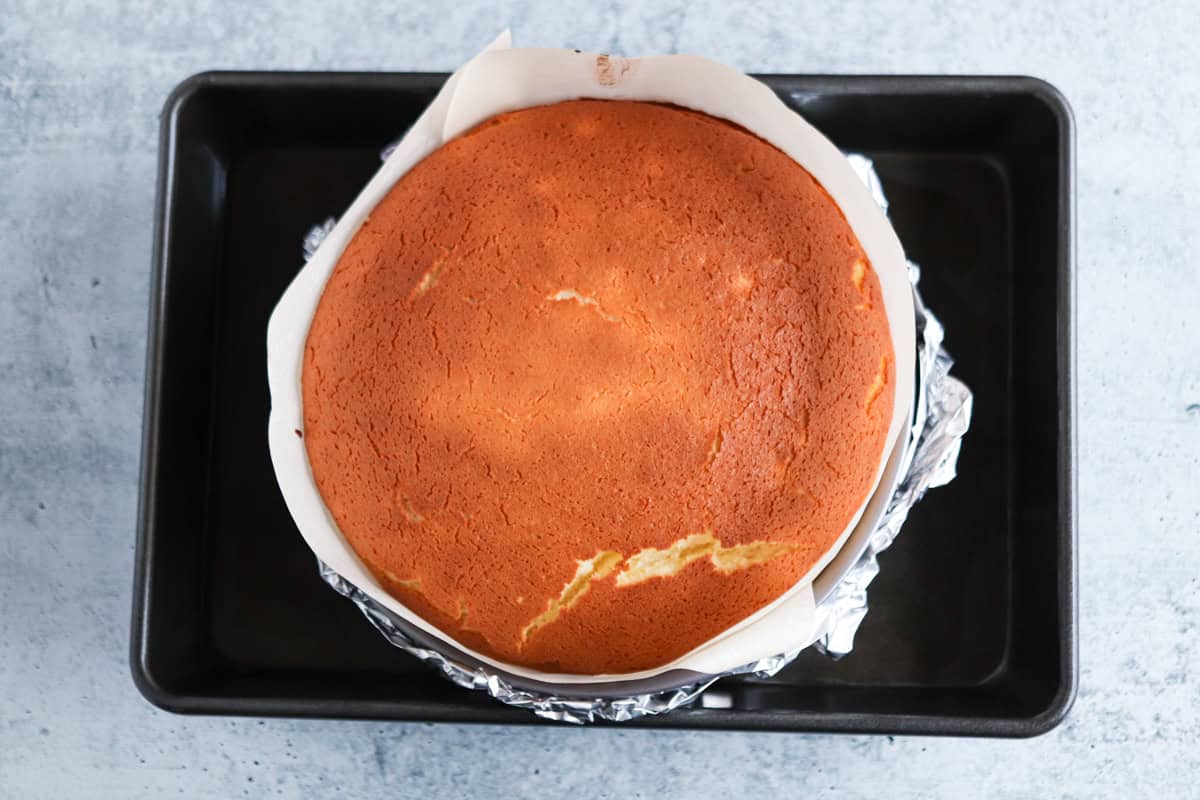A round, golden-brown cake sits in a parchment-lined pan with foil around it, placed inside a black rectangular baking tray on a light gray surface.