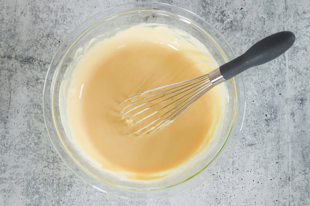 A glass bowl filled with light batter is being whisked with a metal whisk featuring a black handle, placed on a gray textured surface.