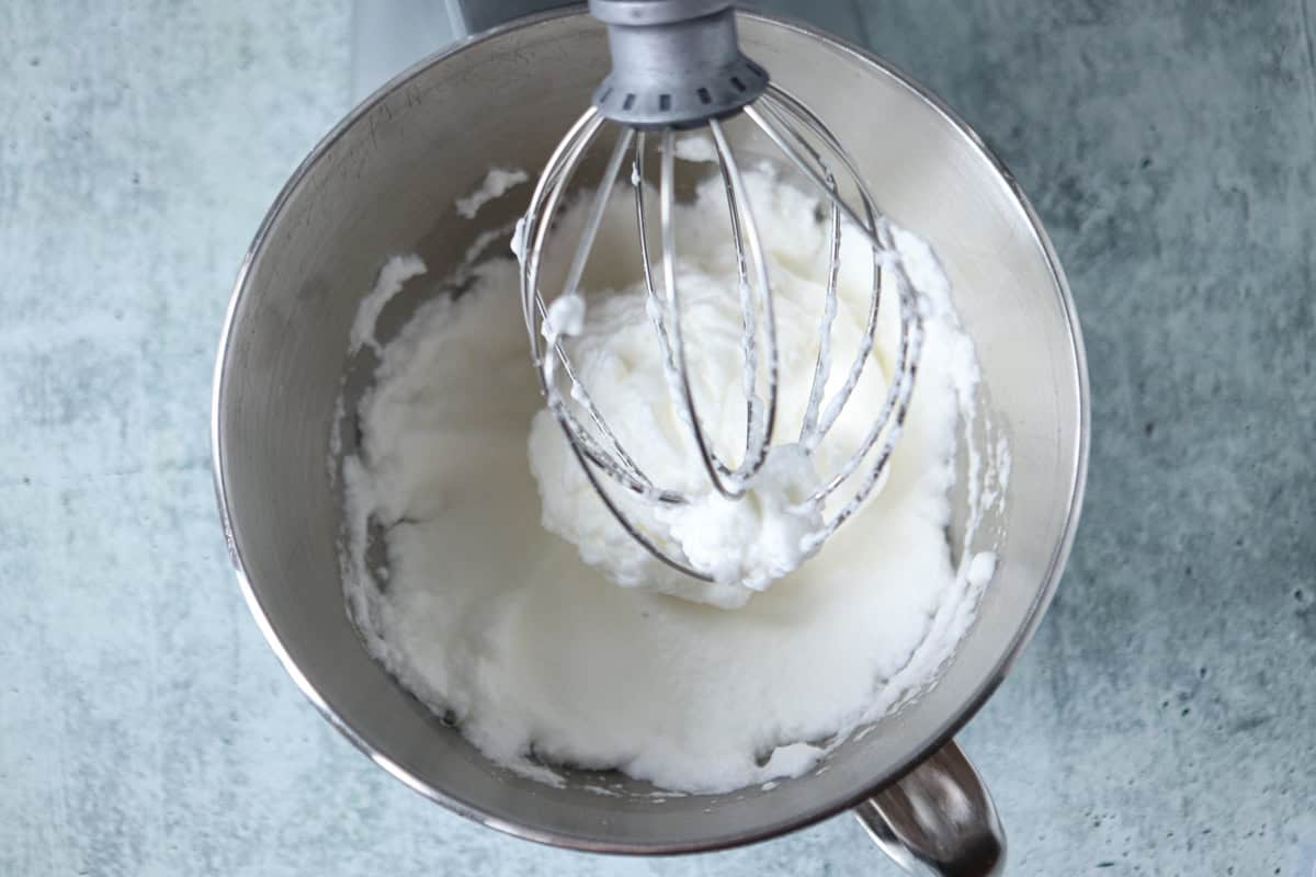 A metal mixing bowl with whipped egg whites and a whisk attachment from a stand mixer, set on a light blue countertop.