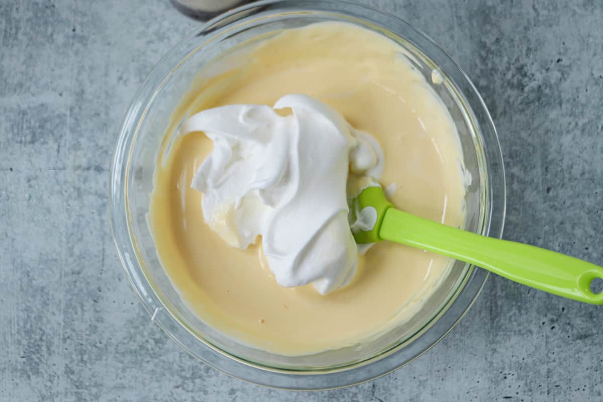 A glass bowl filled with creamy yellow batter, is topped with a dollop of fluffy whipped egg whites. A green spatula rests inside, all set for mixing on the gray textured surface.