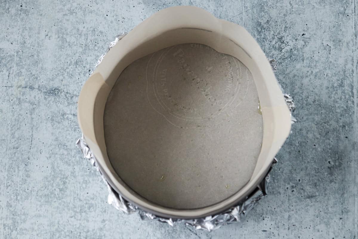 A round baking pan, is lined with parchment paper on the sides and bottom and wrapped in aluminum foil, sitting on a gray textured surface.