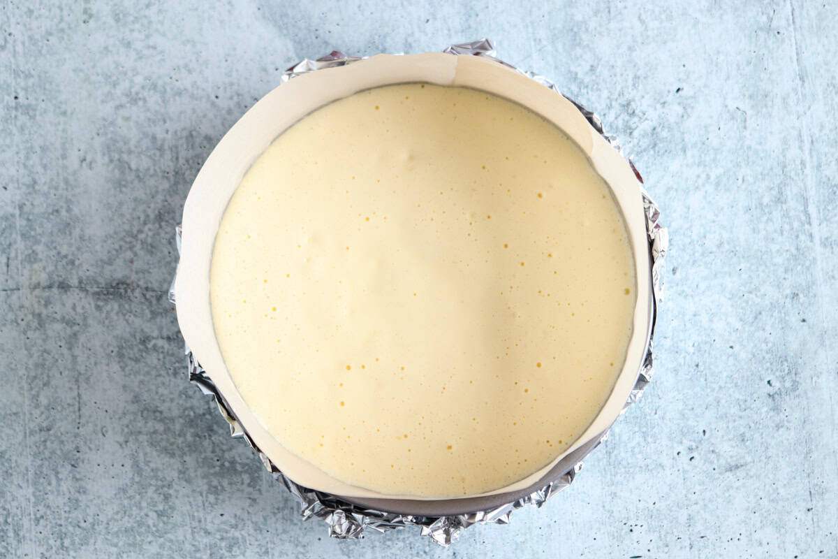 A round cake pan lined with parchment paper and foil, filled with pale yellow, smooth batter, sits on a light gray textured surface.