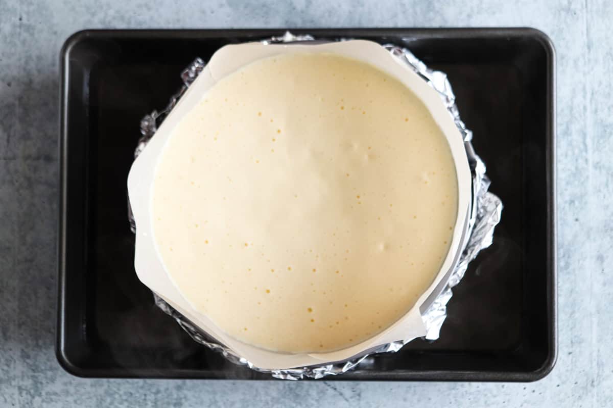 A round cake pan filled with uncooked cheesecake batter, wrapped in foil, sits in a water bath inside a rectangular baking tray.