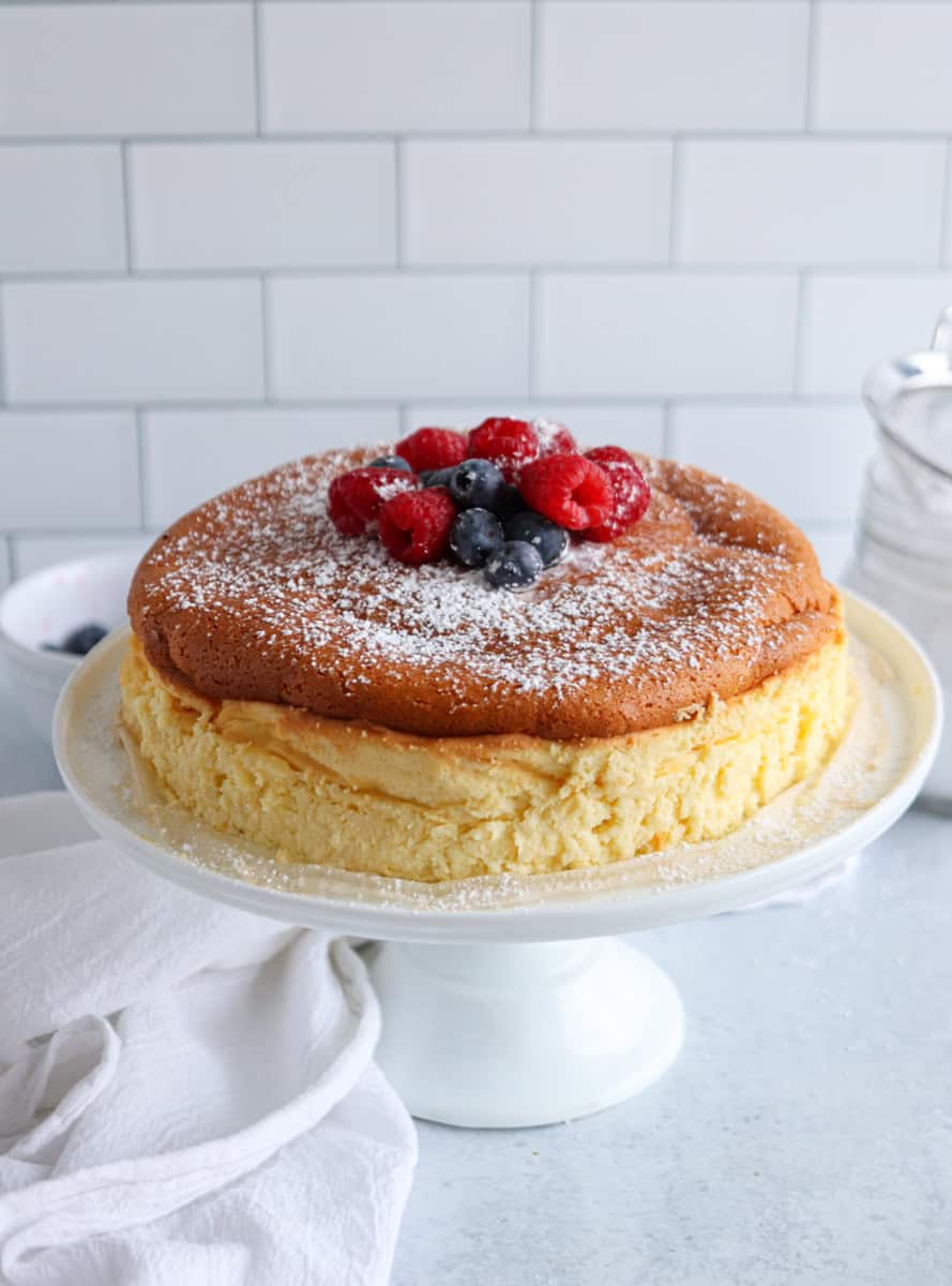 A fluffy, golden-brown skyr cheesecake sits on a white cake stand, topped with powdered sugar and fresh raspberries and blueberries. The background features white subway tiles and a white cloth.