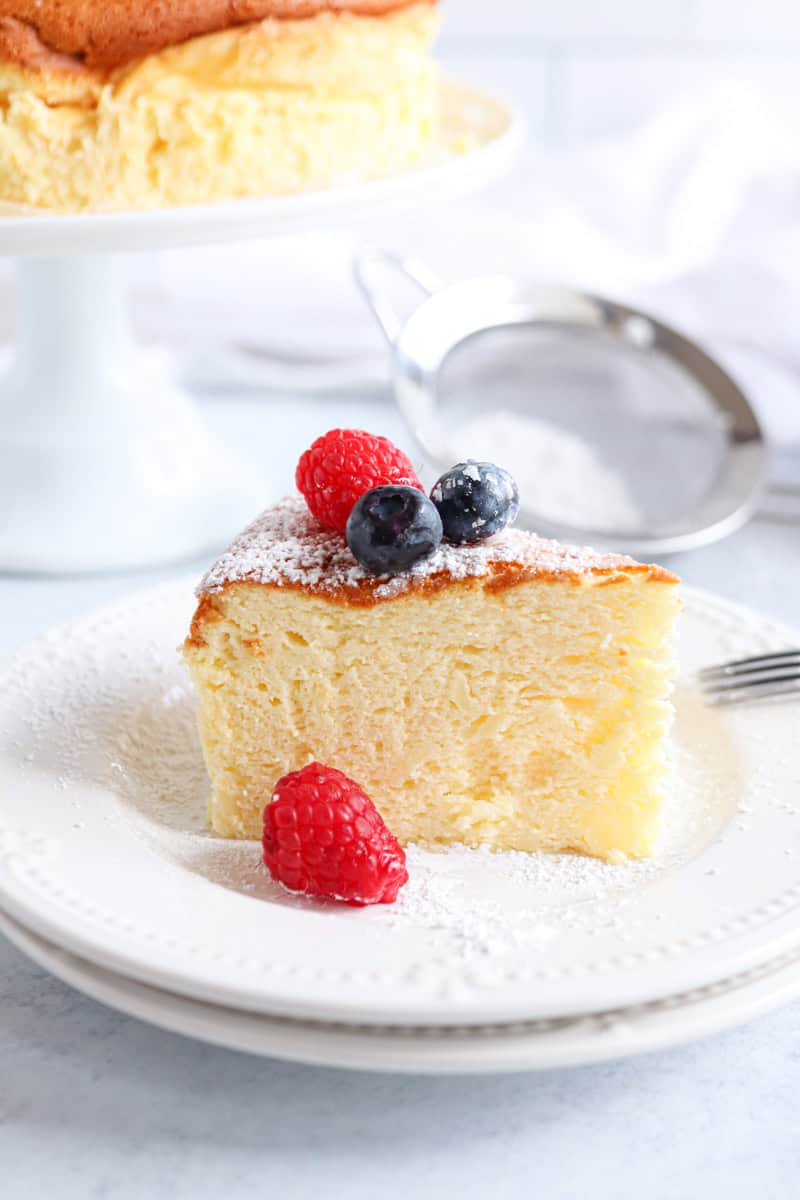 A slice of fluffy skyr cheesecake on a white plate, topped with powdered sugar, fresh raspberries, and blueberries, with a cake stand and metal sifter in the background.