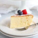 A slice of fluffy cheesecake topped with a raspberry and blueberries, dusted with powdered sugar, sits on a white plate.