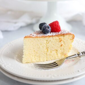 A slice of fluffy cheesecake topped with a raspberry and blueberries, dusted with powdered sugar, sits on a white plate.