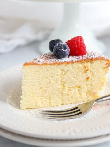 A slice of fluffy cheesecake topped with a raspberry and blueberries, dusted with powdered sugar, sits on a white plate.
