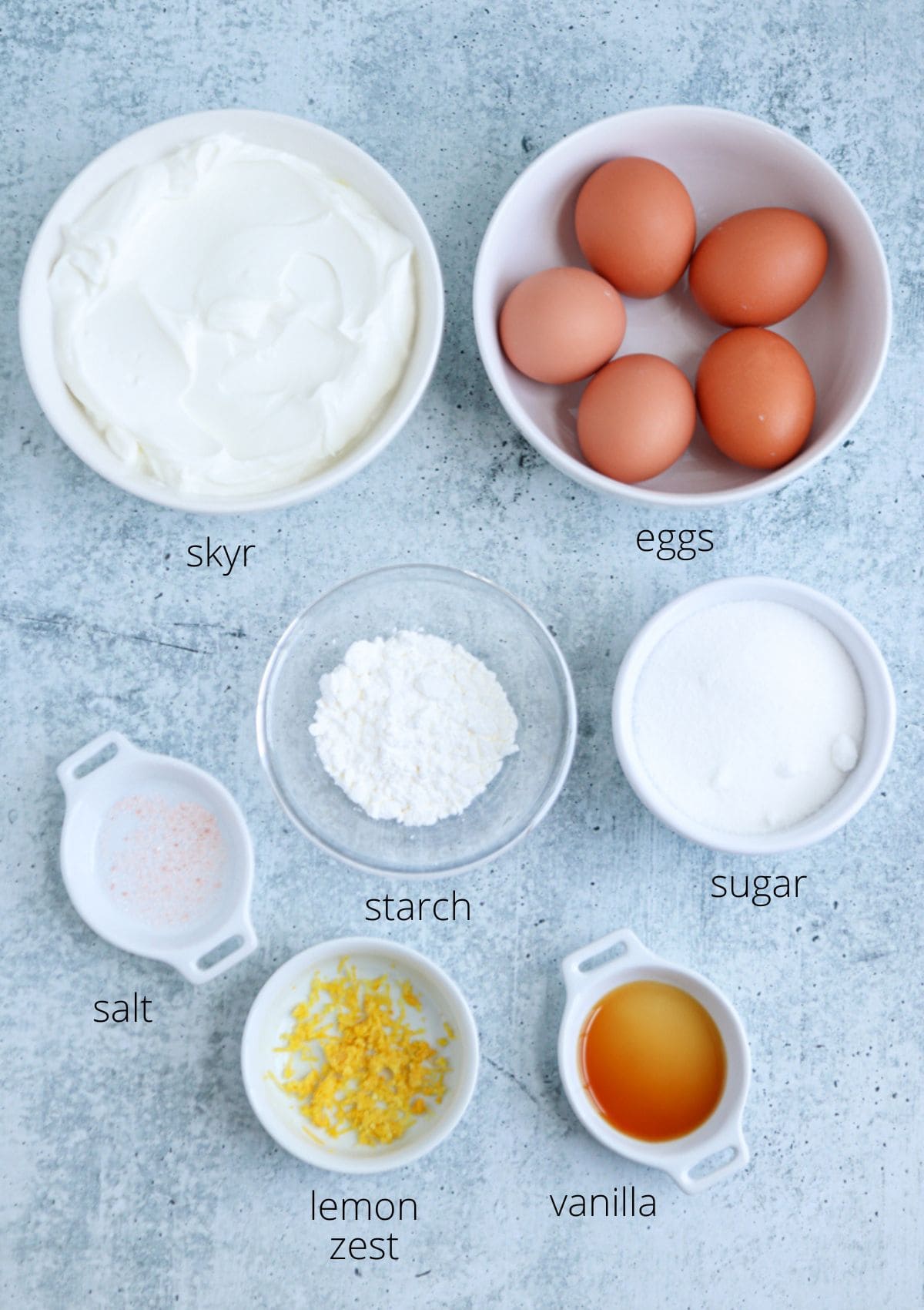 A flat lay of seven bowls containing skyr, eggs, starch, sugar, salt, lemon zest, and vanilla, each labeled and arranged on a light gray surface.