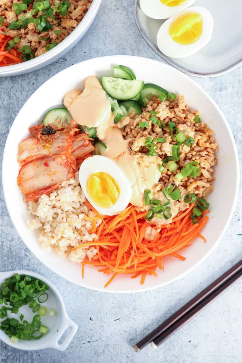 A bowl with sliced cucumber, shredded carrots, kimchi, cooked minced meat, brown rice, half a soft-boiled egg, green onions, and creamy sauce. Chopsticks, green onions, and eggs are nearby on a light surface.