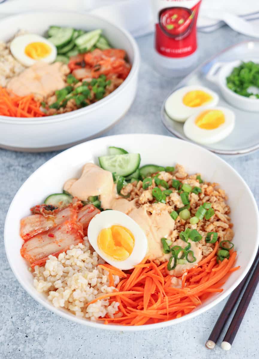 A bowl filled with brown rice, shredded carrots, sliced cucumber, kimchi, chopped green onions, a halved soft-boiled egg, and a drizzle of creamy sauce. Another bowl and sliced eggs are in the background. Chopsticks rest nearby.