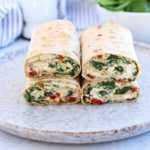 Four halves of a wrap are stacked on a plate, showing layers of spinach, cream cheese, and sun-dried tomatoes inside a tortilla—perfect to pair with high protein chicken alfredo. A bowl of greens and a striped cloth are in the background.