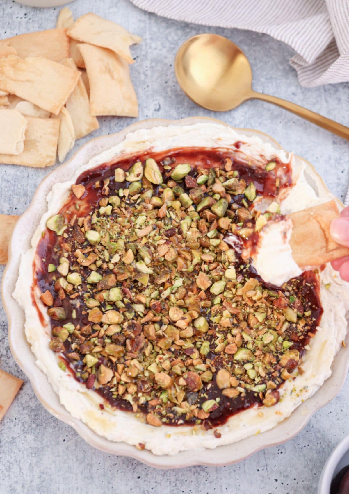 A bowl of creamy boursin dip topped with chopped pistachios and a dark sauce, surrounded by pieces of pita chips. A hand dips a pita chip into the boursin dip. A gold spoon and a napkin are nearby.