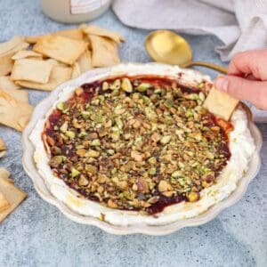 A hand dips pita chip into a dish of whipped Boursin dip topped with jam and chopped pistachios. More pita chips and a gold spoon are nearby on a blue surface.