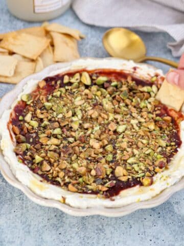 A hand dips pita chip into a dish of whipped Boursin dip topped with jam and chopped pistachios. More pita chips and a gold spoon are nearby on a blue surface.