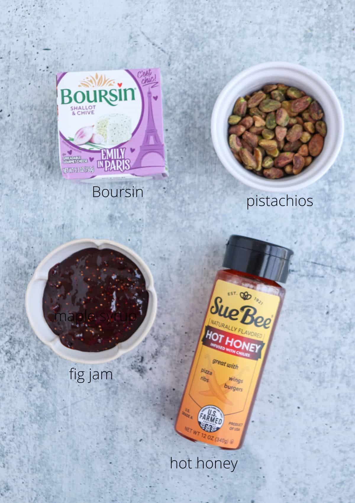 A flat lay of four labeled food items: Boursin shallot & chive cheese perfect for a boursin dip, a small bowl of pistachios, a bowl of fig jam with chili flakes, and a bottle of Sue Bee hot honey, on a gray background.