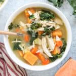 A bowl of chicken and vegetable soup with shredded chicken, kale, carrots, celery, onions, and sweet potatoes sits on a light gray surface next to a striped cloth and a slice of sweet potato.