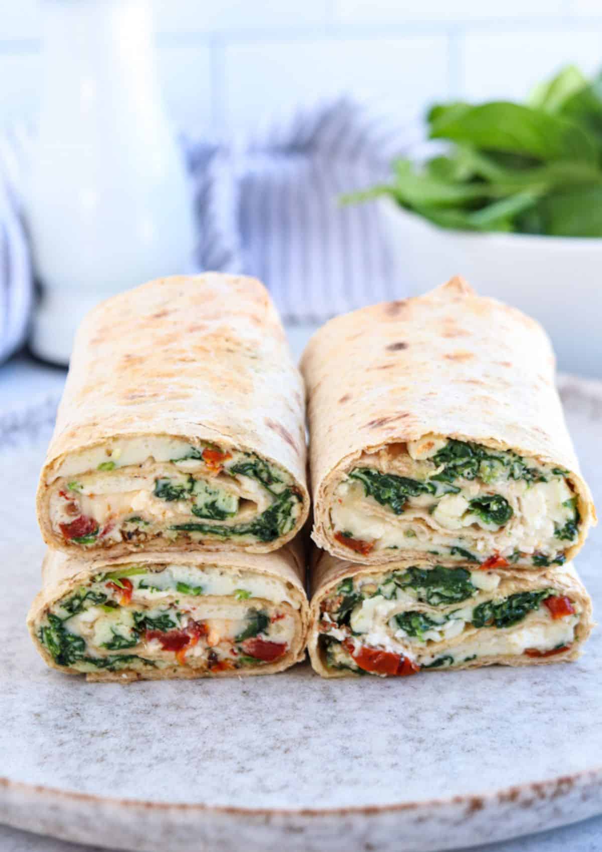 Four halves of veggie wraps filled with spinach, hummus, and chopped red peppers are stacked on a plate. A bowl of fresh spinach and a pitcher blur in the background.