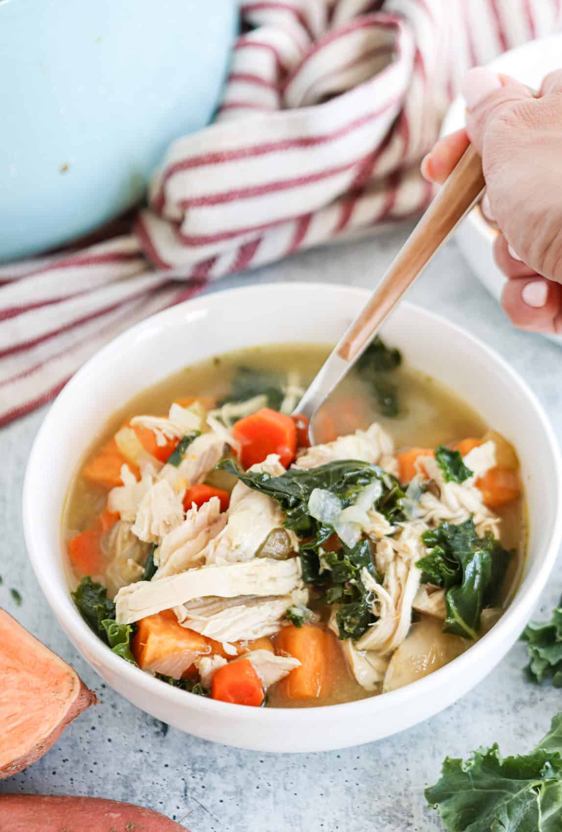 A hand holding a spoon over a bowl of hearty chicken and vegetable soup with carrots, kale, and shredded chicken, set on a table with a striped towel and sweet potato nearby.