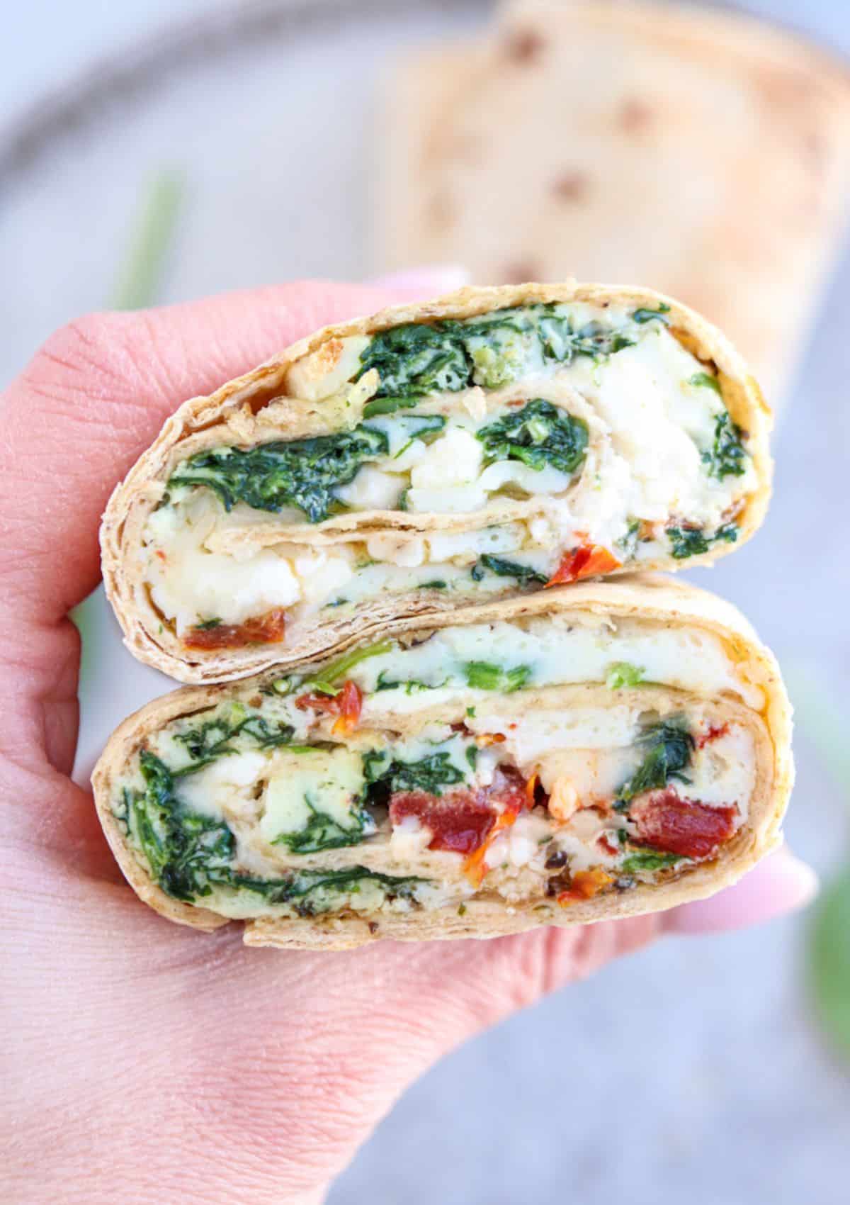 A hand holds a halved wrap filled with spinach, melted cheese, eggs, sun-dried tomato, showing the colorful layered ingredients inside.