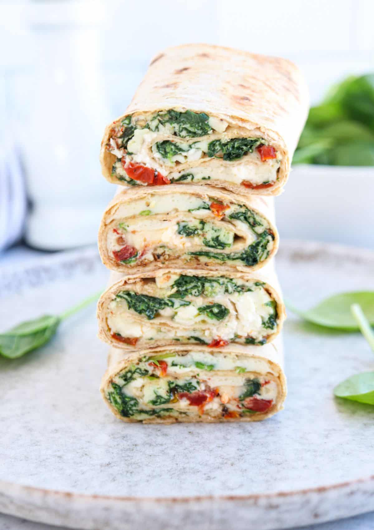 Four halves of a spinach and cheese wrap with red bell peppers are stacked on a plate, showing the colorful filling inside the tortilla. Fresh spinach leaves are scattered around as garnish.