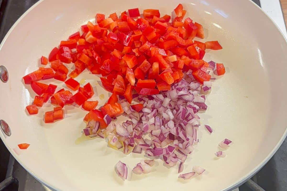 A white frying pan on a stove with chopped red bell pepper and diced red onion pieces, ready to be cooked for a savory cottage cheese egg bake.