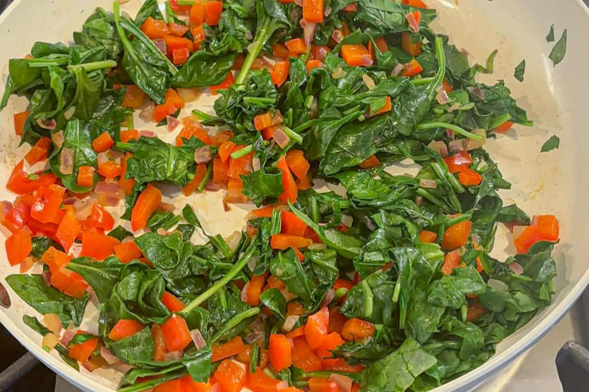 Fresh spinach and diced red bell peppers sautéing in a white skillet on a stovetop, perfect as a flavorful base for your next cottage cheese egg bake.