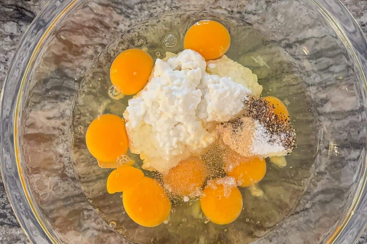 A clear glass bowl contains six raw eggs, cottage cheese, and a mixture of seasonings—ingredients ready to be mixed for a delicious cottage cheese egg bake—placed on a gray marbled countertop.