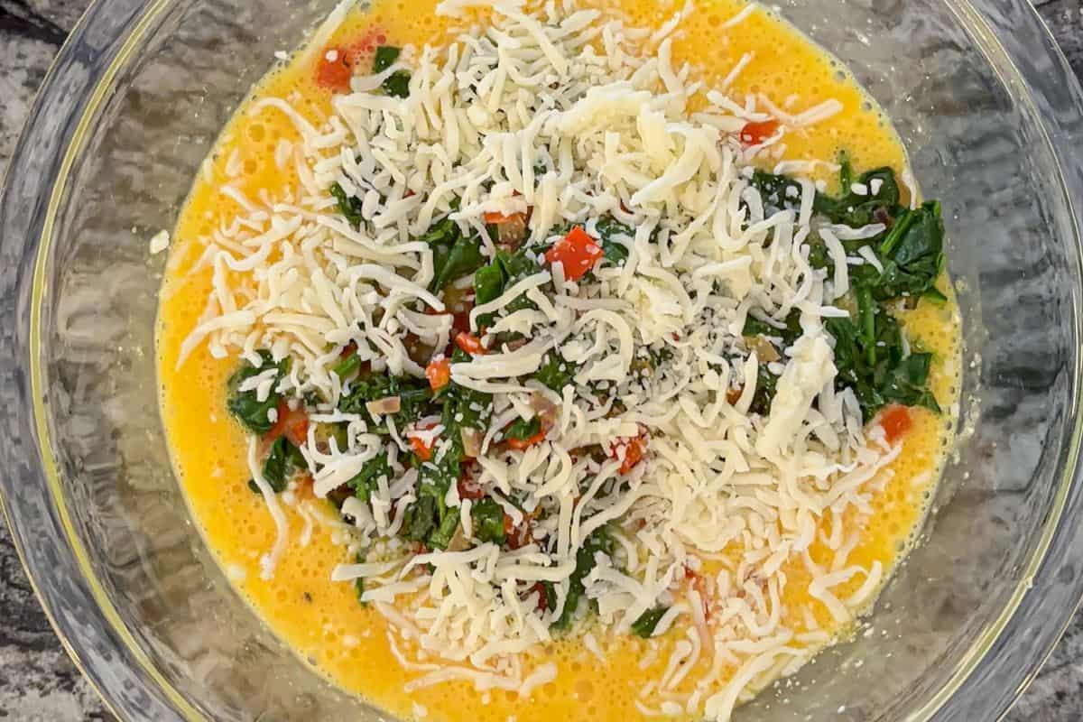A glass bowl filled with beaten eggs, shredded cheese, chopped spinach, and diced red bell peppers, perfect for creating a flavorful cottage cheese egg bake.