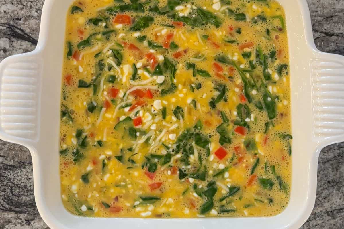 A white square baking dish filled with a colorful cottage cheese egg bake featuring chopped spinach, red bell peppers, and shredded cheese sits on a gray marble countertop.