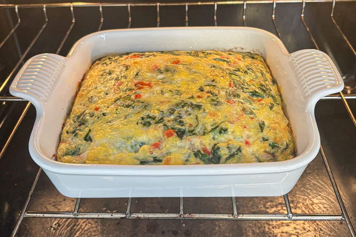 A rectangular white baking dish with a cottage cheese egg bake filled with spinach, red peppers, and cheese sits on an oven rack. The casserole is puffed up and lightly browned on top.
