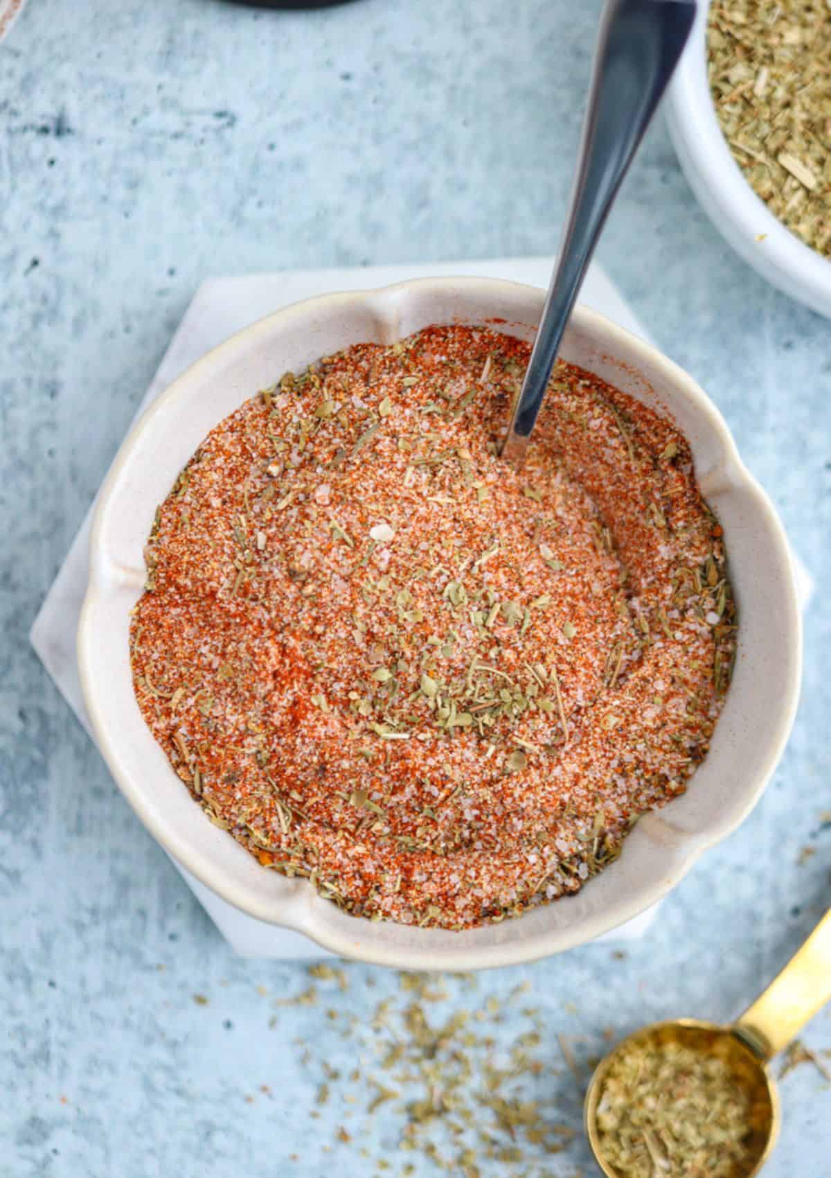 A bowl filled with a mixed dry seasoning blend, featuring red, green, and white specks—perfect for making chicken—with a spoon resting inside. A gold measuring spoon and another bowl of herbs are nearby on a light blue surface.