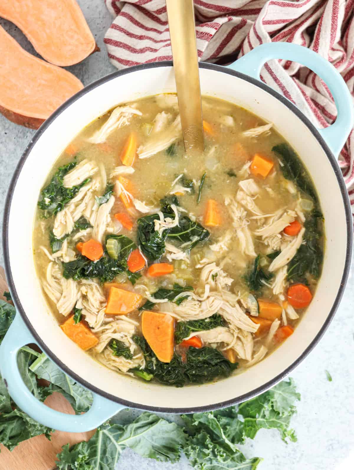A pot of hearty chicken kale soup with shredded chicken, kale, carrots, sweet potatoes, and celery, with a gold ladle inside. Striped cloth and fresh kale are nearby.