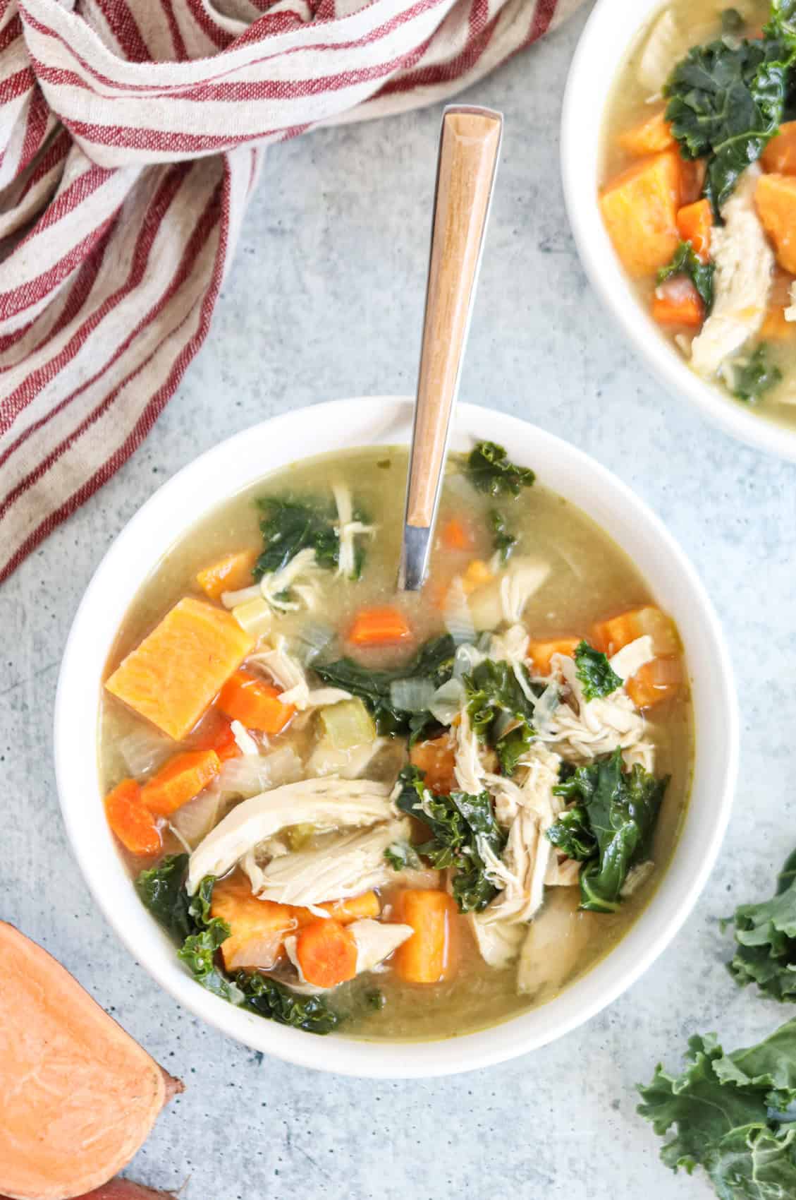 A bowl of chicken soup with shredded chicken, kale, carrots, and sweet potatoes. A spoon rests inside, with a striped cloth and a partial bowl of soup visible beside it.