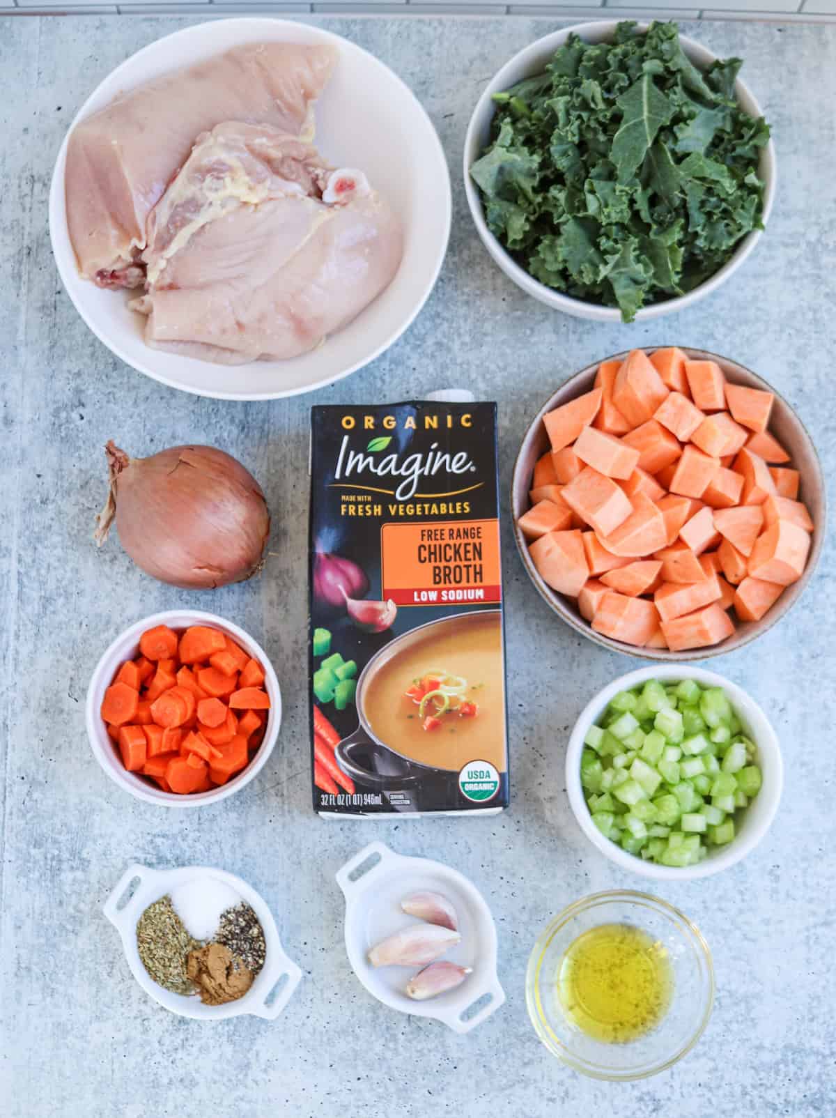 Bowls of raw chicken breast, chopped kale, diced sweet potatoes, sliced carrots, celery, shallot, garlic cloves, olive oil, spices, and a carton of organic chicken broth are arranged on a light surface.