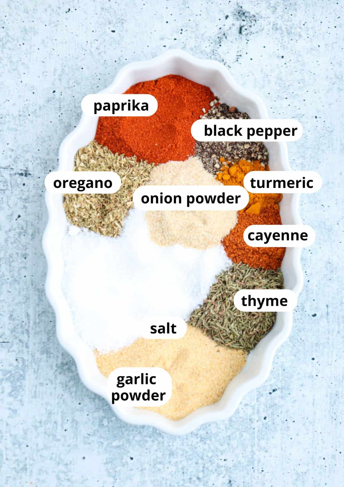 An oval white dish holds piles of various spices—perfect for seasoning a homemade boursin dip—each labeled: paprika, black pepper, turmeric, cayenne, thyme, oregano, onion powder, salt, and garlic powder on a light gray surface.