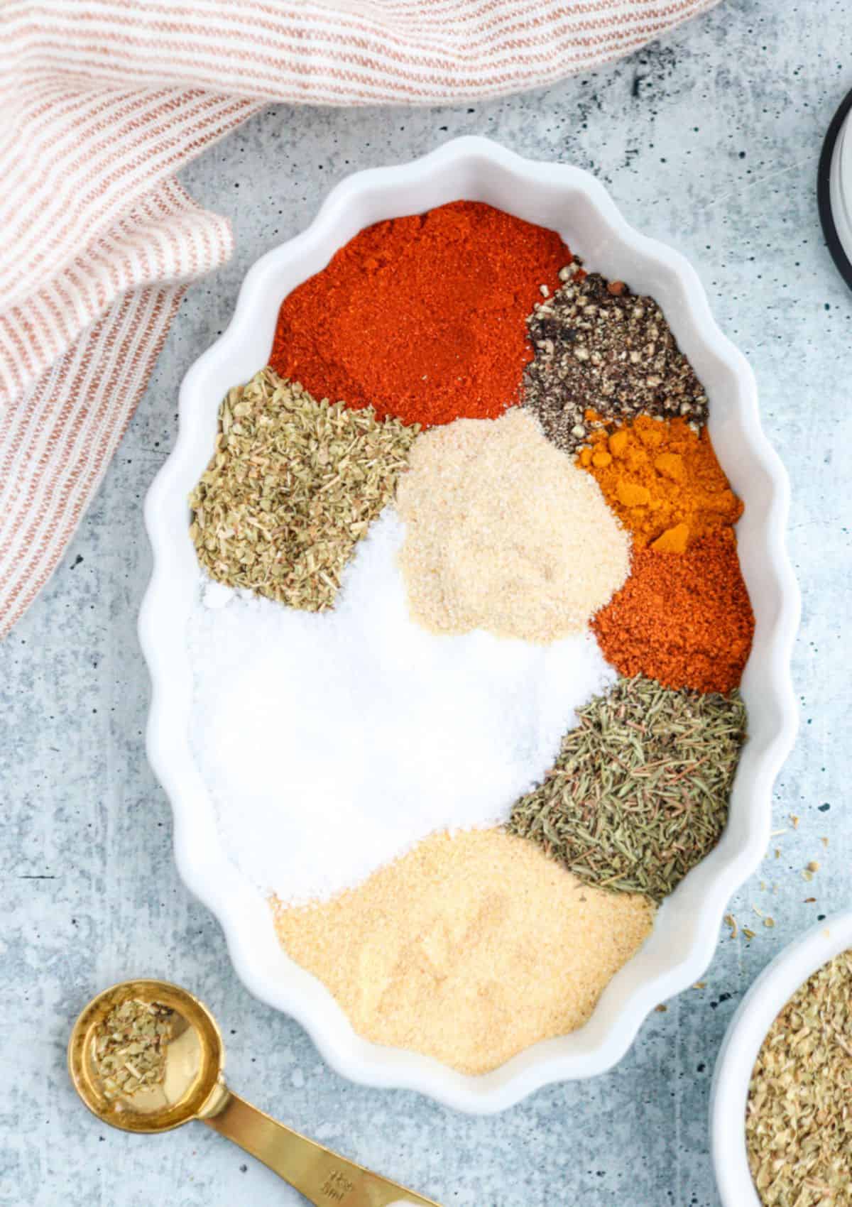 An oval white dish holds small piles of various spices for boursin dip, including paprika, black pepper, dried herbs, salt, onion powder, turmeric, and garlic powder. A gold measuring spoon with herbs sits nearby on a textured gray surface.