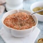 A spoon holds a scoop of mixed dried spices and herbs for boursin dip over a white bowl filled with more of the seasoning blend, set on a light surface with additional spices in the background.